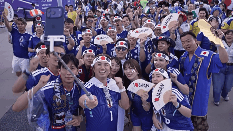 A imagem ilustra torcedores do Japão tirando uma selfie do lado de fora de um estádio.