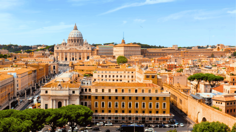 Vista panorâmica da Cidade do Vaticano, com destaque para a Basílica de São Pedro ao centro, cercada por edifícios históricos, muralhas do Vaticano e árvores sob céu azul.