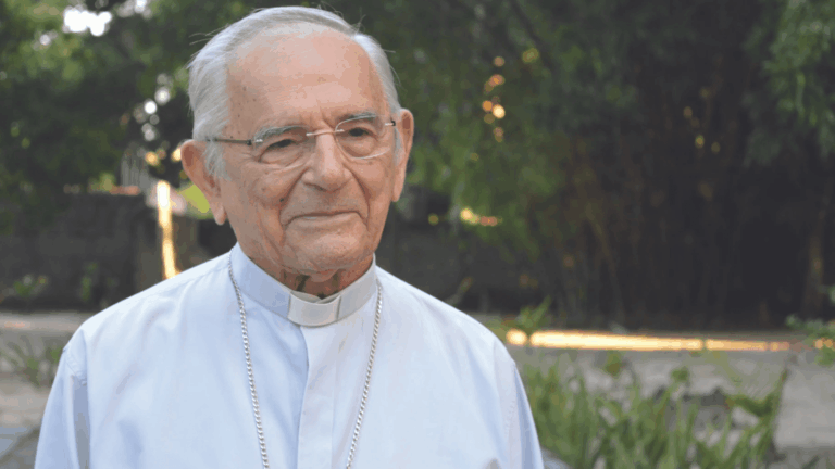Dom Heitor usa uma camisa de manga longa azul claro com colarinho clerical branco (clérgima) e óculos. Ao seu fundo há uma vegetação