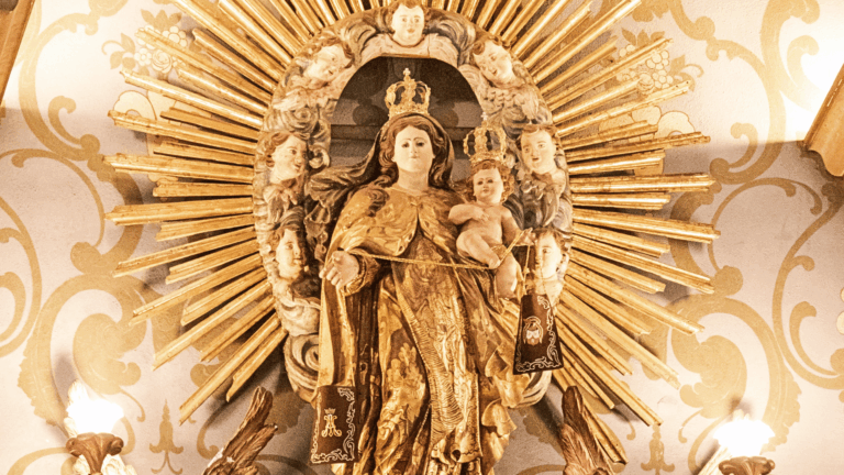 Imagem de Nossa Senhora do Carmo com o Menino Jesus ao colo, ambos coroados, tendo ao fundo um resplendor dourado cercado por anjos. A Virgem segura o escapulário, símbolo da devoção carmelita.