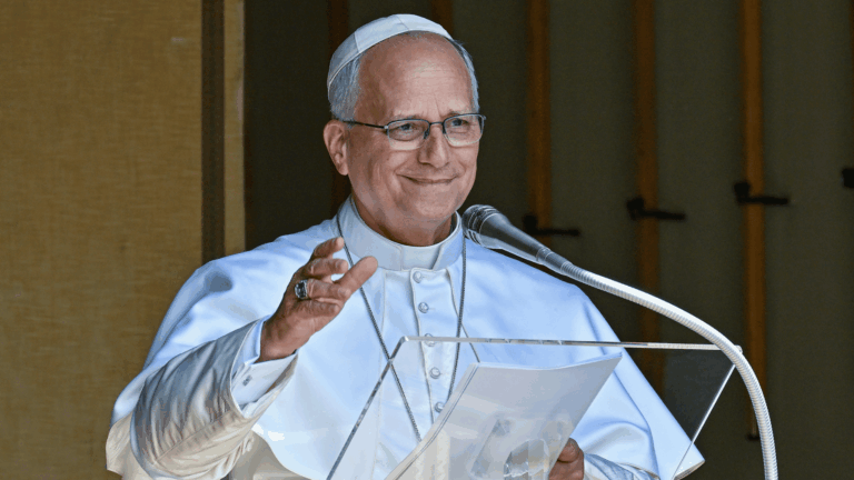 O Papa Leão XIV aparece com suas habituais vestes brancas diante de um púlpito com microfone. Ele acena com a mão direita