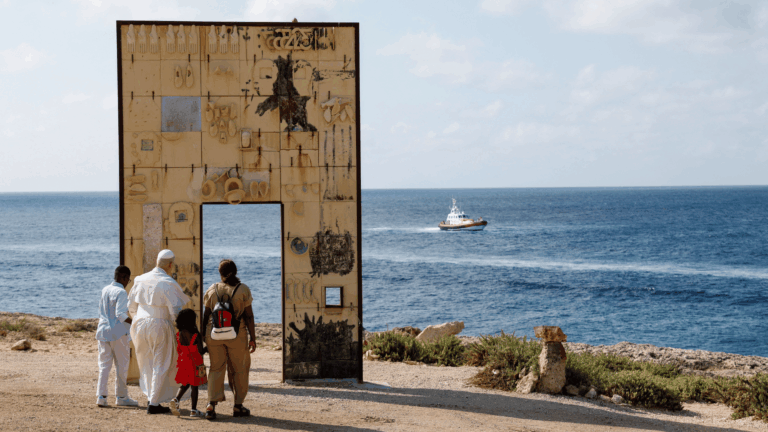 A imagem ilustra o Papa Leão XIV, com suas habituais vestes pontifícias, junto a uma família de migrantes. Eles visitam um monumento intitulado "Porta da Europa", que consiste em um grande pórtico com vista para o Mar Mediterrâneo, presente no fundo da imagem.