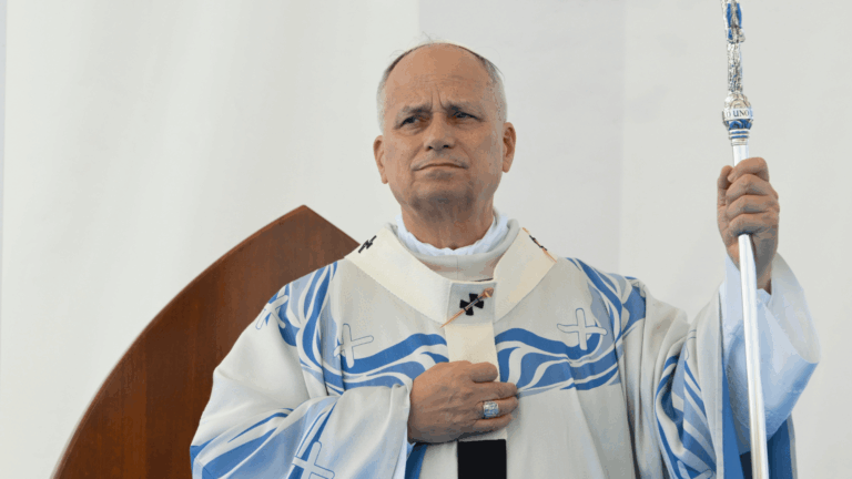 A imagem ilustra o Papa Leão XIV, com expressão séria, vestida com paramentos litúrgicos brancos com detalhes azuis, durante a celebração da Missa