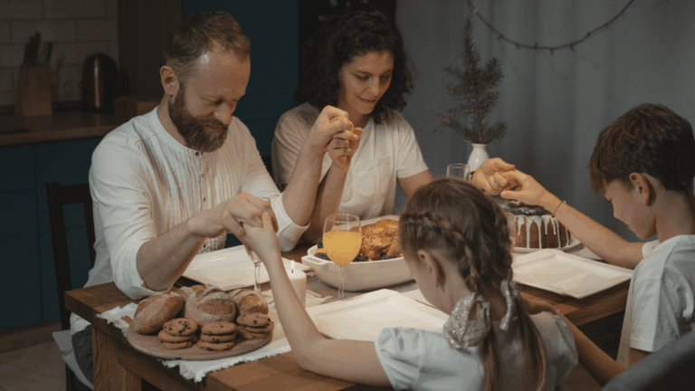 A foto mostra uma família reunida à mesa: há um homem, uma mulher e duas crianças (um menino e uma menina). Todos estão de mãos dadas, como se estivesse rezando antes da refeição. A mesa possui travessas com biscoitos, bolo e taças com sucos.