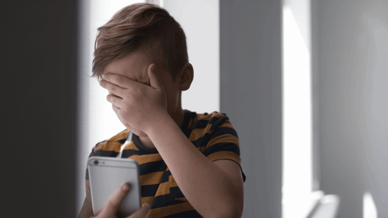 Criança cobre o rosto enquanto segura um celular, representando os perigos da internet para crianças.