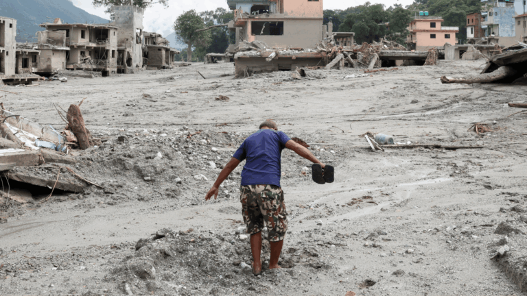 A imagem mostra um homem caminhando pela lama em meio a um cenário de destruição após inundações no Nepal