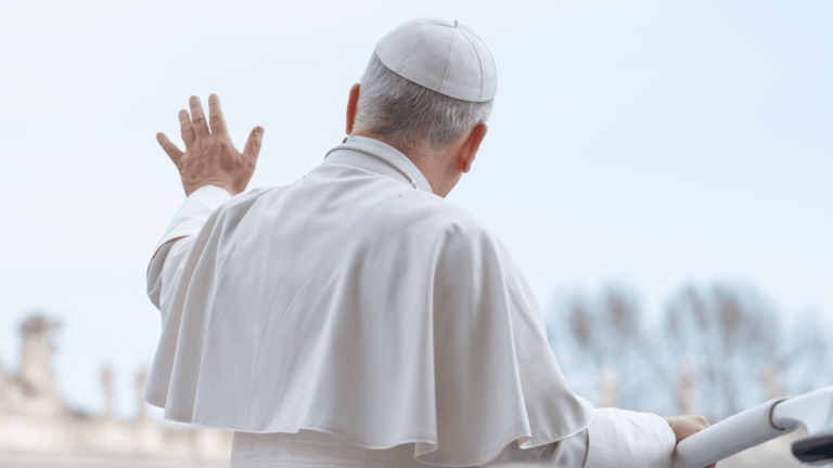 A foto mostra o Papa Leão XIV, de costas, acenando. Ele veste suas habituais vestes brancas