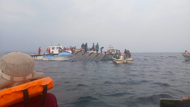 A imagem mostra uma embarcação virada no mar e outra embarcação da equipe de resgate que trabalham com coletes laranjas