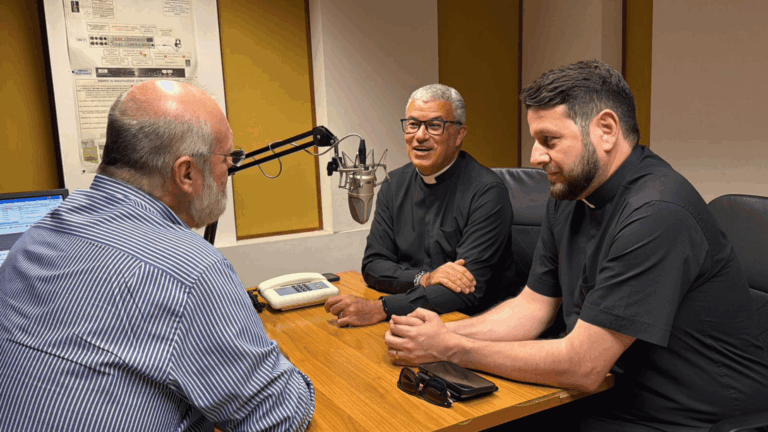 A imagem mostra os padre Roger Luis e Donizete Heleno sentados a uma mesa em frente ao jornalista Silvonei José