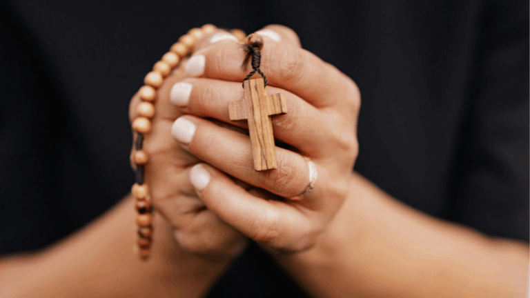 Mãos unidas em oração seguram um terço de contas de madeira com crucifixo, em imagem de destaque para a prática da oração e da devoção cristã.