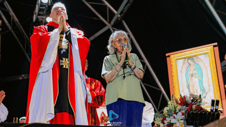A imagem mostra a tia Lolita, fundadora do Hallel, em pé, de camiseta verde e calça azul, com as mãos unidas em posição de oração e segurando um microfone. AO lado dela, está o padre Marcelo Rossi, com vestes litúrgicas, na mesma posição. Eles estão em pé em um palco.