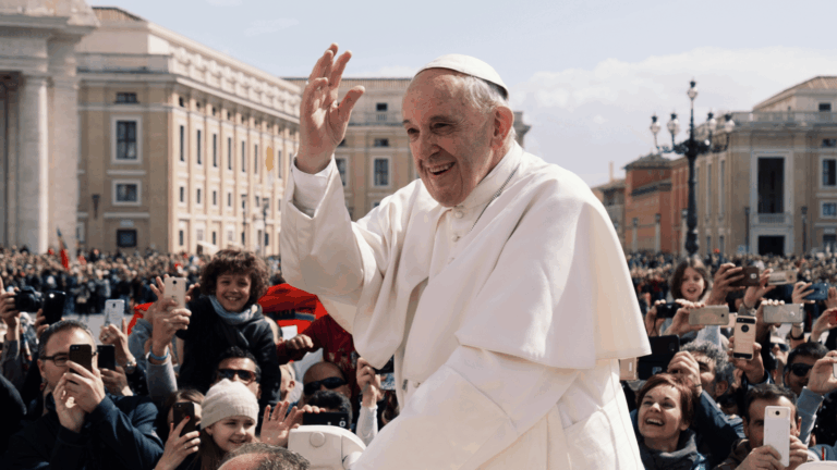 Papa Francisco, com as tradicionais vestes brancas, passa pela Praça São Pedro de papamóvel enquanto acena aos fiéis.