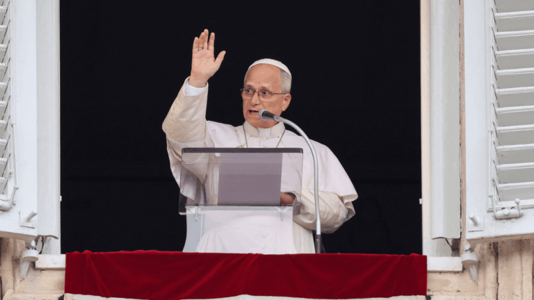 A imagem mostra o Papa Leão XIV na janela do palácio apostólico vaticano. Ele acena aos fiéis e fala ao microfone.