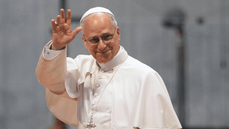 A imagem ilustra o Papa Leão XIV, vestido com suas habituais vestes pontifícias brancas, com o braço direito erguido, acenando.
