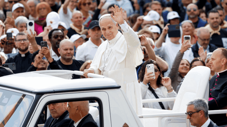 A imagem mostra o PApa Leão XIV com suas habituais vestes brancas. Ele está em pé no papamóvel e acena aos fiéis que estão ao redor na Praça São Pedro