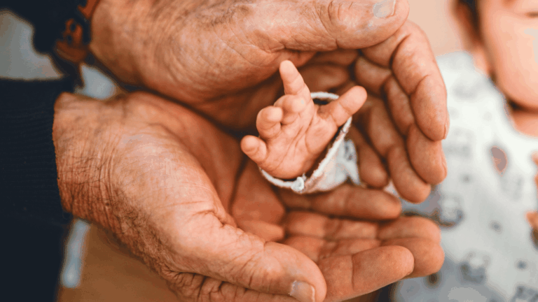 Mãos de um adulto envolvem delicadamente a pequena mão de um bebê.